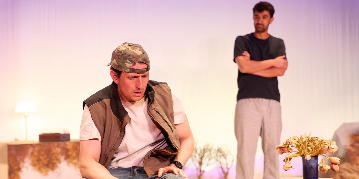 The background is lilac and white. There is a person sitting down. Behind them another person can be seen standing, the image of them is blurry.