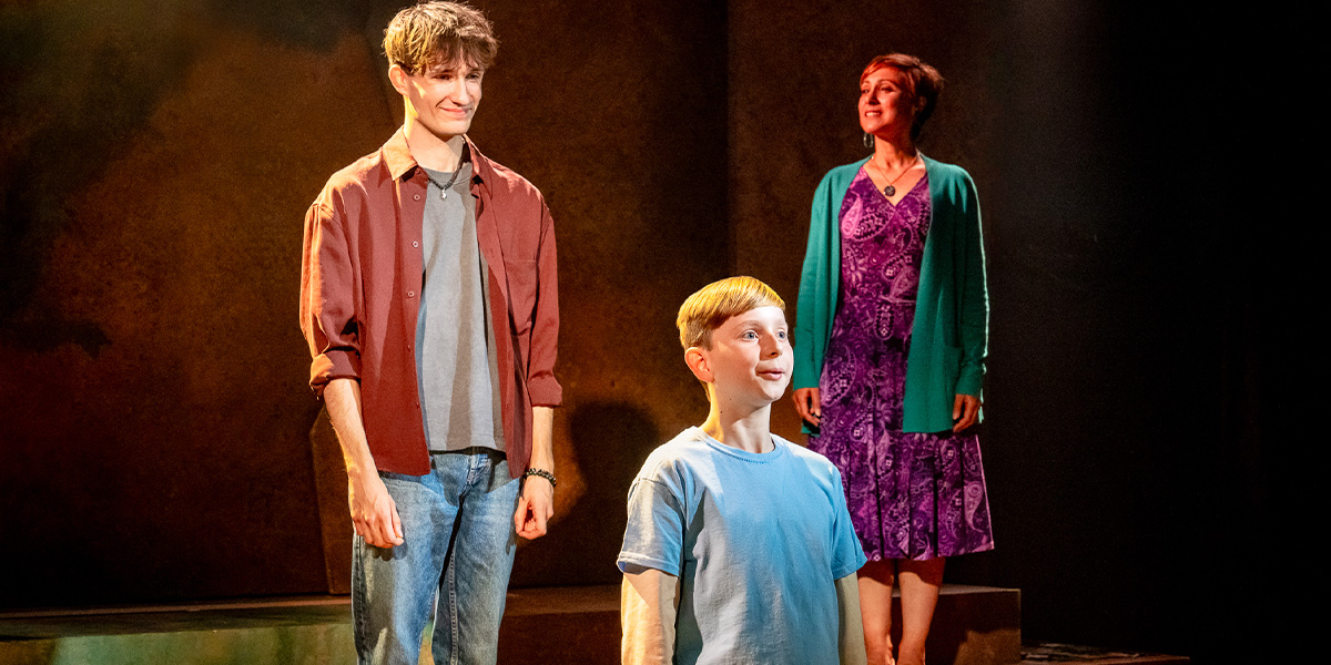 Three actors performing on stage, with one in the foreground looking up, and two in the background observing. They appear in a scene involving emotional interaction.
