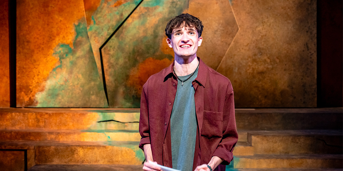 Person standing on stage with an abstract backdrop, smiling and wearing a maroon jacket over a blue shirt.