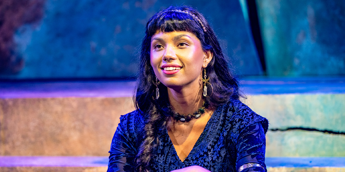 Person with long dark hair and bangs, wearing a blue dress and a necklace, sitting and smiling onstage with theatrical lighting.