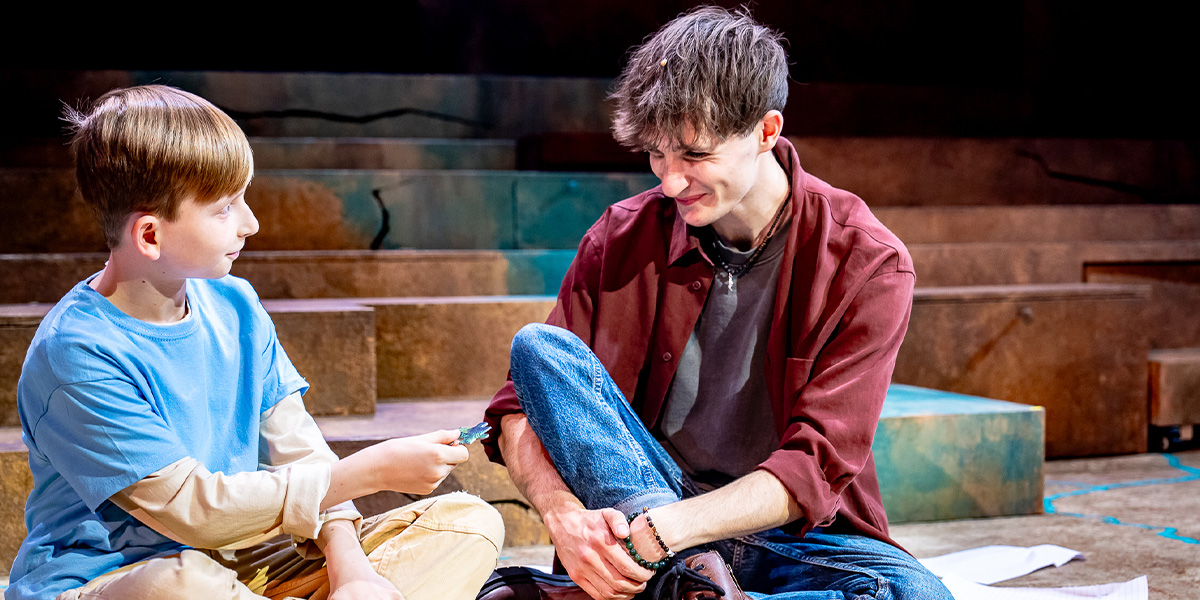 Two actors are on stage, with one person handing a small object to the other, both smiling and engaging in a conversation.