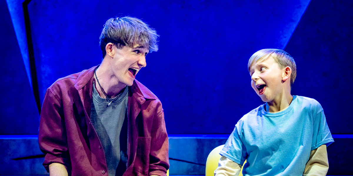 Two actors on stage portraying a joyful scene in a theatrical performance, one wearing a red jacket and the other in a blue shirt, both smiling and facing each other against a vibrant blue backdrop.