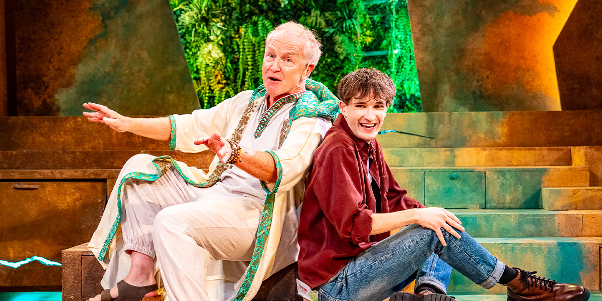 Two actors performing on stage in a theatrical production, one dressed in vibrant, traditional garb and seated, gesturing expressively, and the other in casual modern clothing, sitting on steps and smiling. The backdrop features ornate, stone-like set designs.