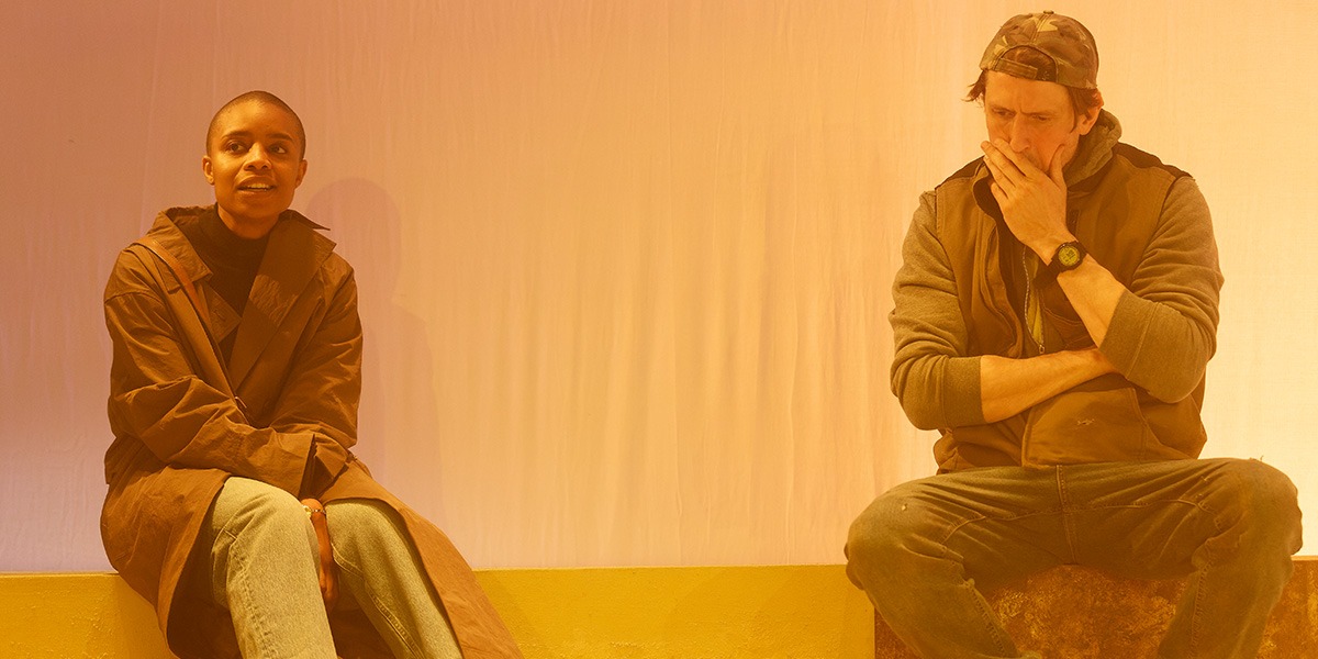 The background is pale yellow. There are two people sitting on a sofa. The person on the left is smiling. The person on the right has their hand to their mouth and looks as if they are deep in thought.