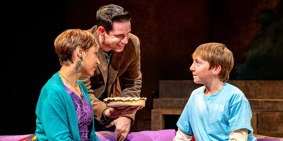 Three individuals are engaged in a cheerful encounter in a theatrical setting, one presenting a pie to the others, who are seated by a bed.