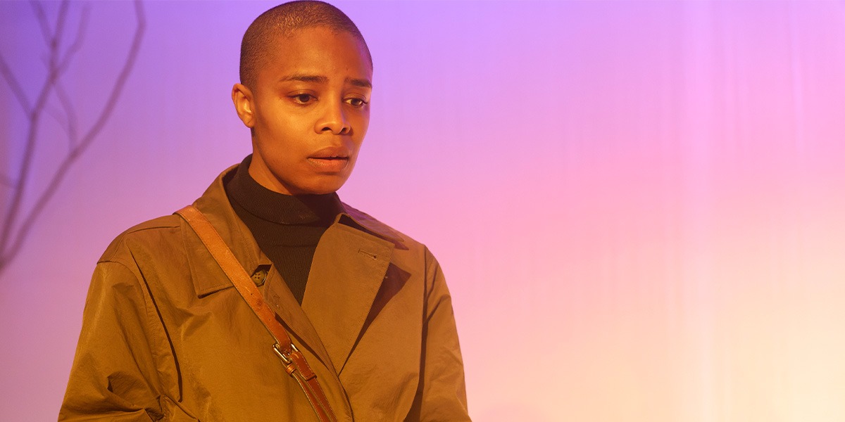 The background is pink and lilac. There is a person standing on the left who is wearing a brown jacket. They are looking down.