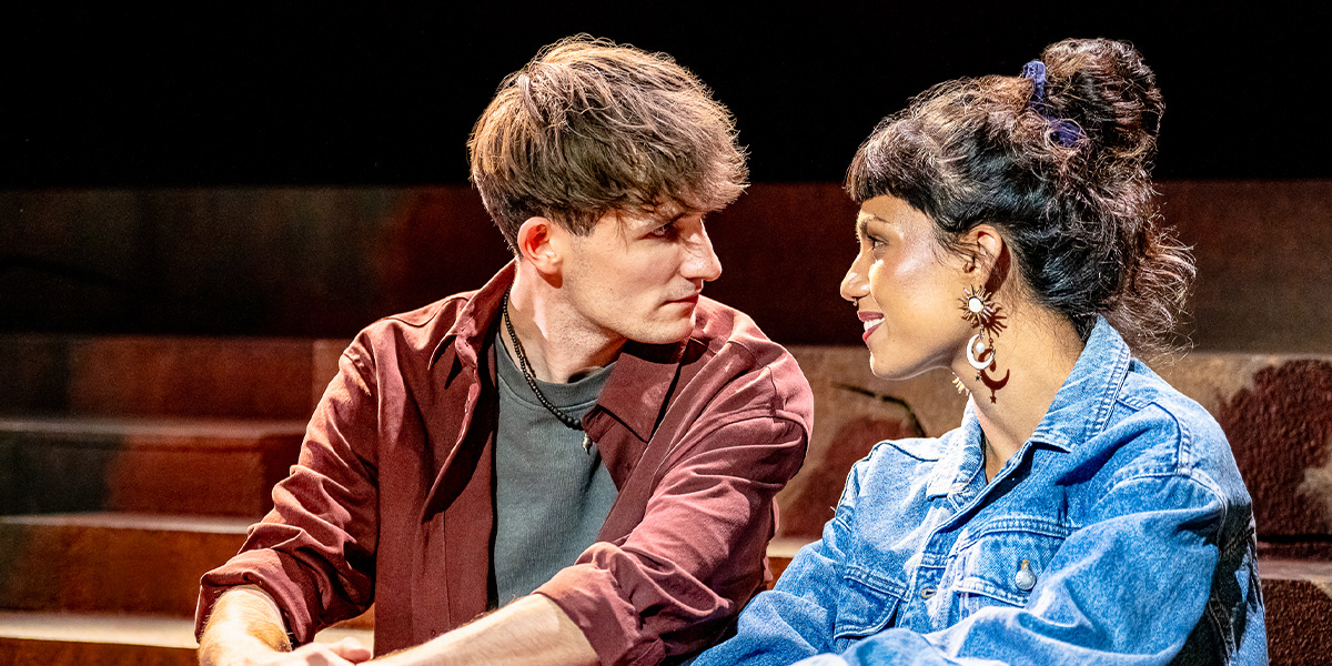 Two people sitting close together on a stage, engaged in an intimate conversation. The person on the left is wearing a maroon jacket and the person on the right is wearing a denim jacket and has their hair styled in a bun. They appear to be actors performing in a theatrical production.
