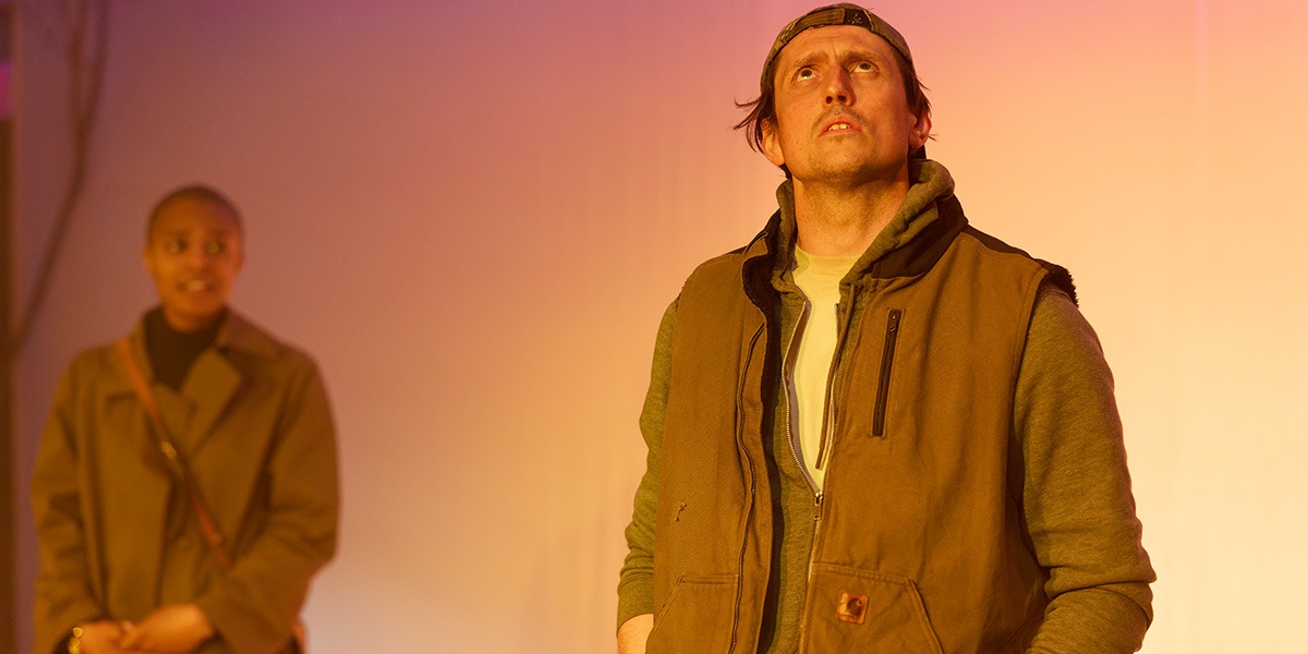 The background is pink and yellow. There is a person standing to the right who is wearing a brown jacket. They are looking up. A person can be seen in the background. The image of this person is blurred.