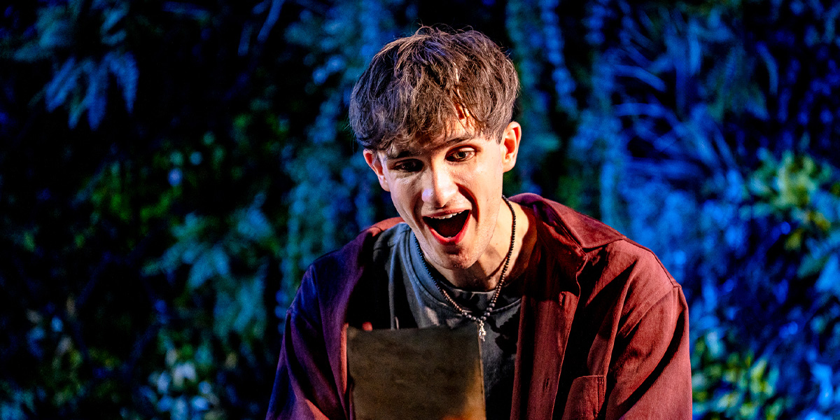 A performer looks excitedly at a map during a theatrical performance, with a backdrop of dense, dark foliage.