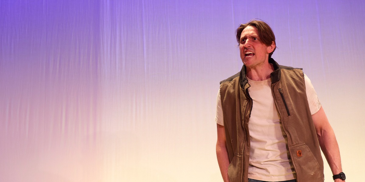 The background is lilac. There is a person standing on the right who is wearing a white t-shirt and a brown gilet. The person looks angry.