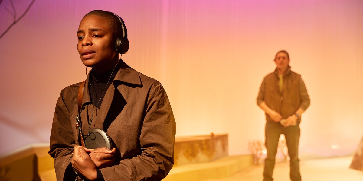 The background is pink and yellow. There is a person standing on the right who is wearing a brown coat and has black headphones over their ears. There is a person standing in the background, the image of this person has been blurred.