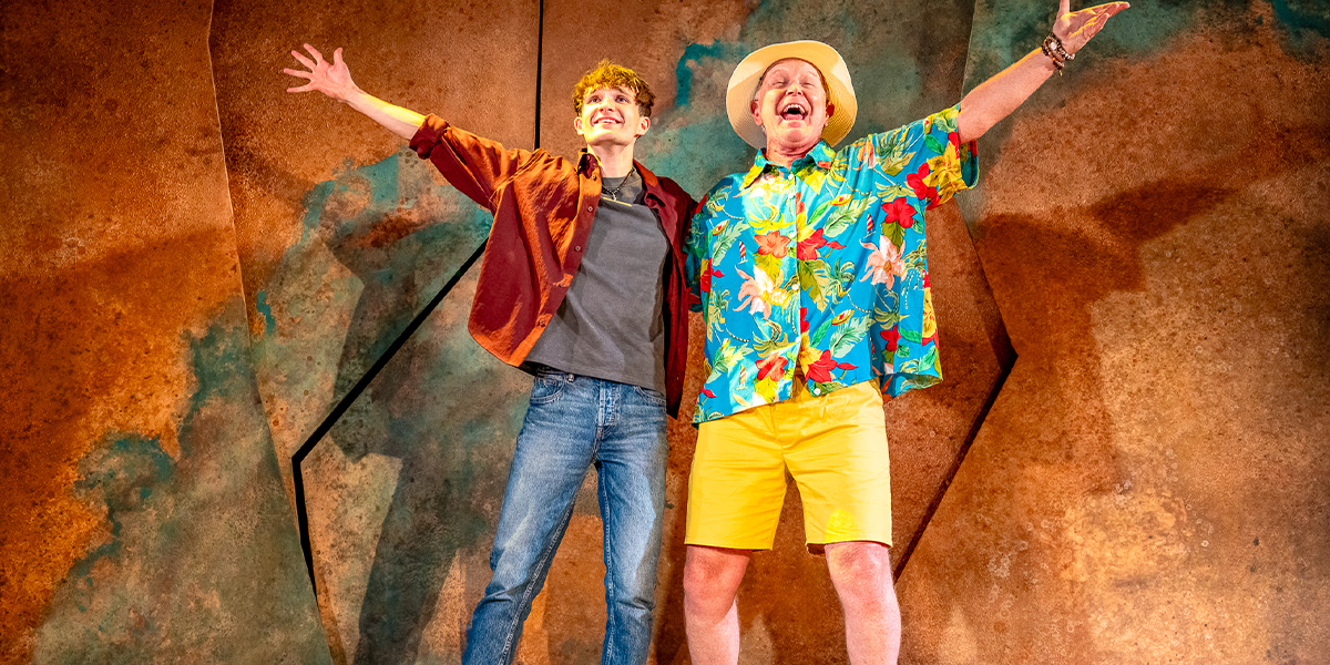 Two actors on stage, one in a floral shirt and hat, and the other in a brown jacket, both smiling and arms raised in a joyful pose.