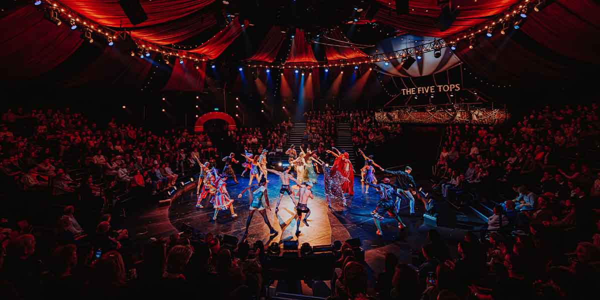 A vibrant circus performance featuring dancers in colourful costumes on stage, with an audience in the background.
