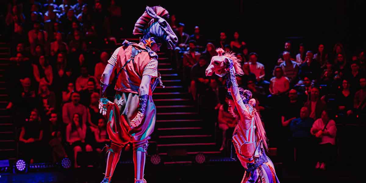 Two performers in vibrant costumes, one with a horse head, entertain an audience in a darkened theatre.