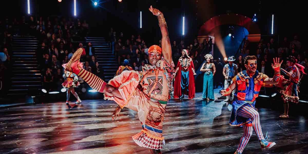 A performer in vibrant costume strikes a dynamic pose on stage, surrounded by fellow cast members.