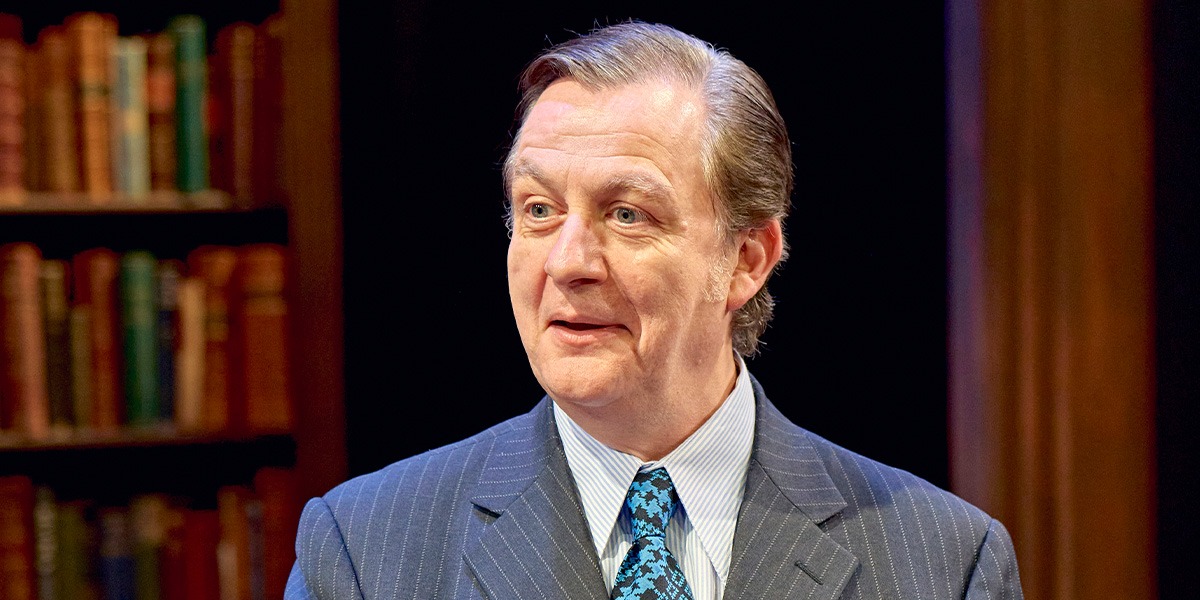 Alan Cox (Tony Crosland) is wearing a grey, striped suit jacket, a white shirt and a blue checked tie and is smiling.