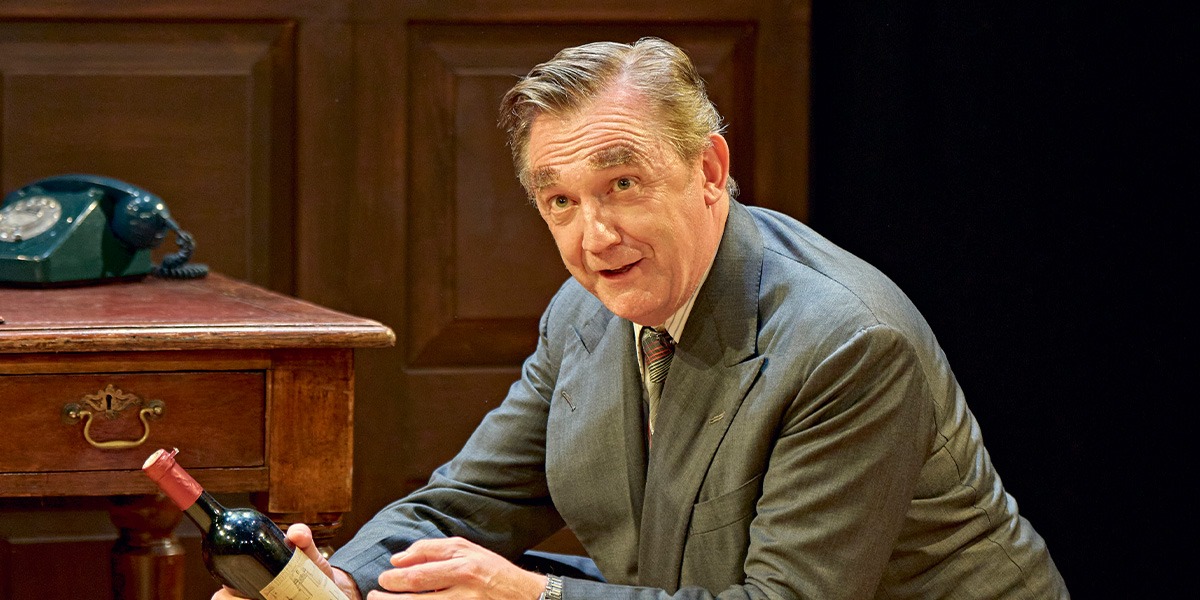 Colin Tierney (Denis Healey) is wearing a grey suit and is sitting at a desk looking amused.