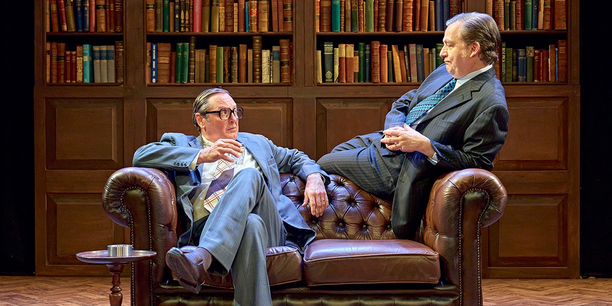Hywell Morgan (Roy Jenkins) and Alan Cox (Tony Crosland) are sitting on a brown leather sofa. In the background a large, full bookcase can be seen.