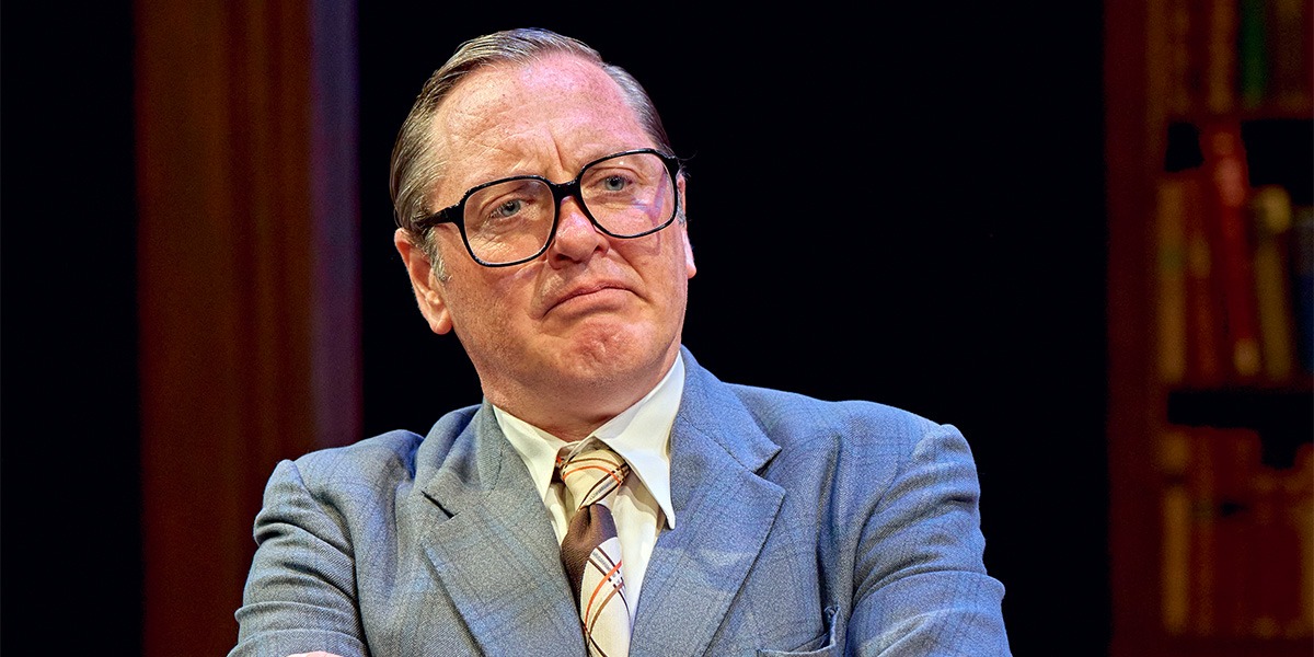 Hywell Morgan (Roy Jenkins) is wearing a grey suit jacket, white shirt, an ivory and brown tie and glasses. Hywell Morgan (Roy Jenkins) appears unhappy