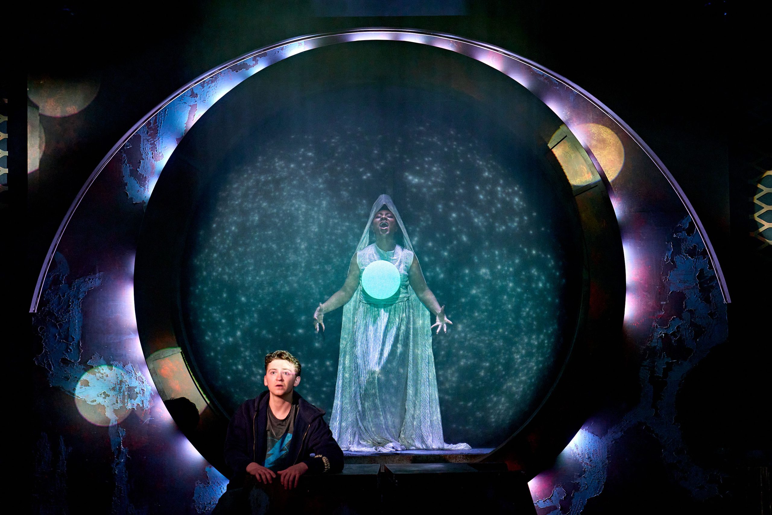 A circular, metallic portal with glowing purple and blue accents serves as the central stage element. Inside the portal, a performer is dramatically posed, dressed in a flowing, shimmery white costume with a translucent veil covering their head. They are holding their arms wide, conveying power or mystical influence, with a glowing orb or circular prop placed near their chest. The backdrop behind them resembles a star-filled, cosmic scene, enhancing the ethereal and celestial atmosphere. In the foreground, another performer sits on the floor, looking up with a mixture of awe and fear, emphasizing the magical or intense nature of the moment. The overall mood is otherworldly and intense, suggesting a dramatic revelation or encounter.