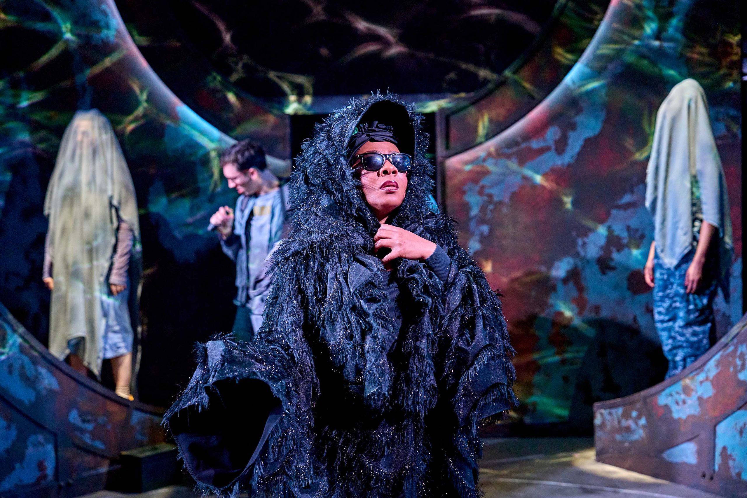 The central figure is dressed in a dramatic, textured, black shaggy costume with a large hood and sunglasses, exuding a powerful and mysterious presence. They are confidently posing or gesturing, drawing attention to themselves. In the background, other performers are visible, some partially covered in translucent fabric that resembles veils or ethereal cloaks, contributing to the surreal and mystical aesthetic. The set design includes curved structures with abstract, colorful patterns. The overall atmosphere is magical and otherworldly, suggesting a fantasy or mythological theme.