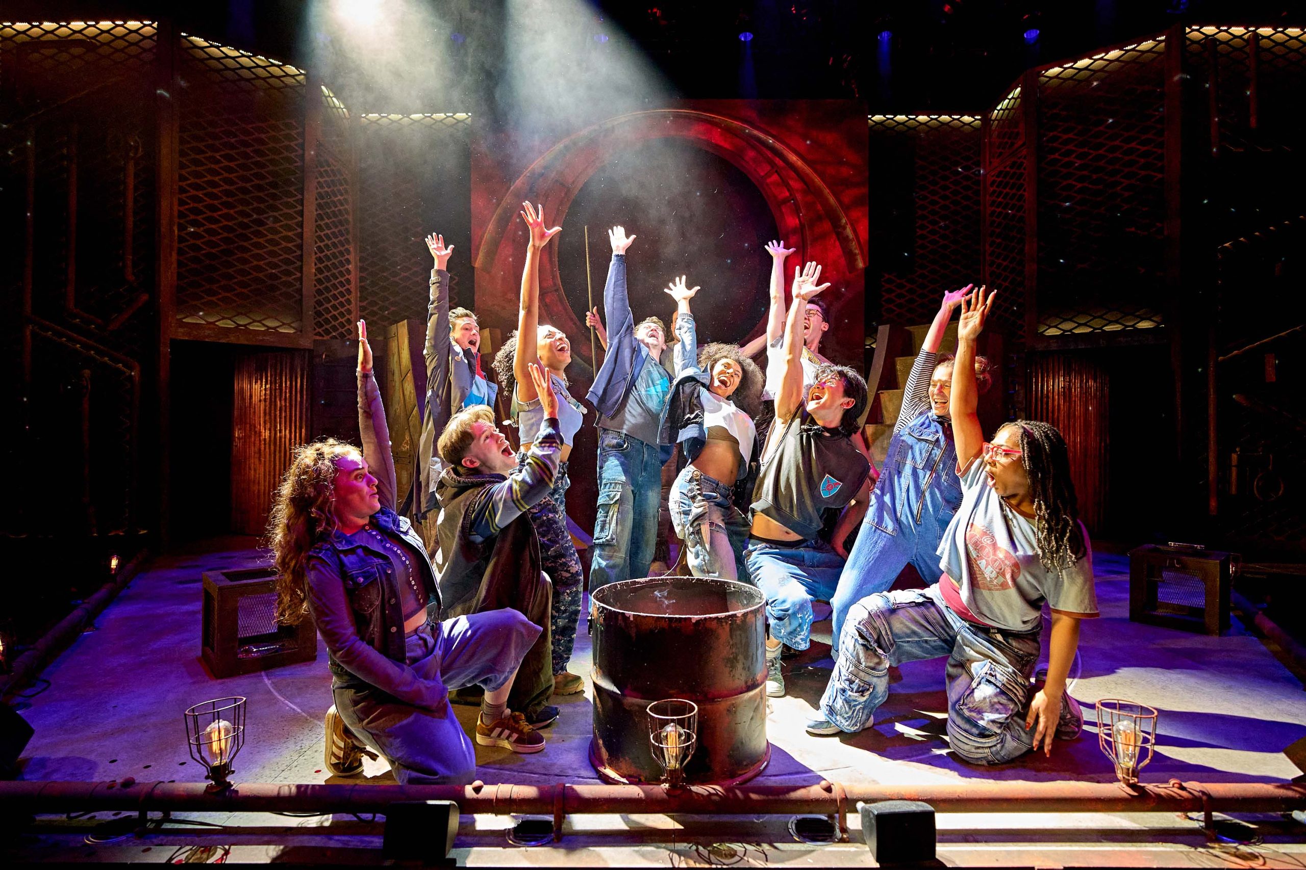 The company of The Lightning Thief are kneeling on stage with one arm raised to the sky.
