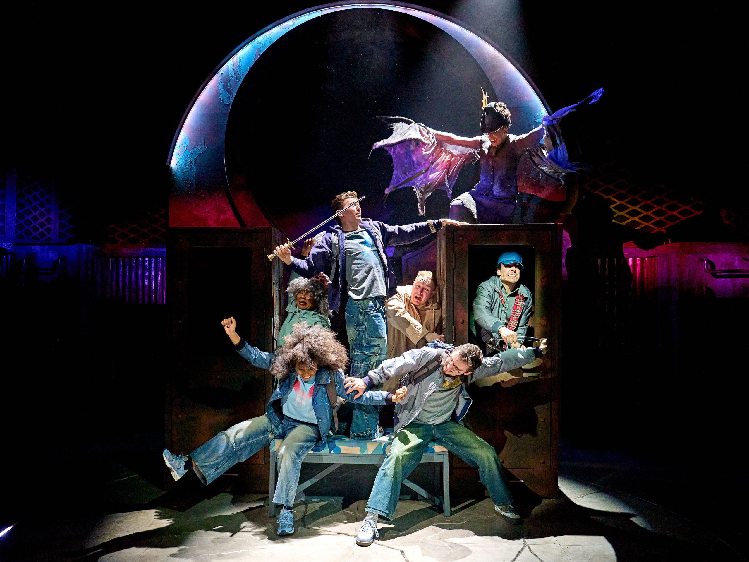 The company of The Lightning Thief sit on steps on stage. At the top of the steps is a large, brown, wooden box. On the box sits a mythical creature with pink and purple flowing wings.