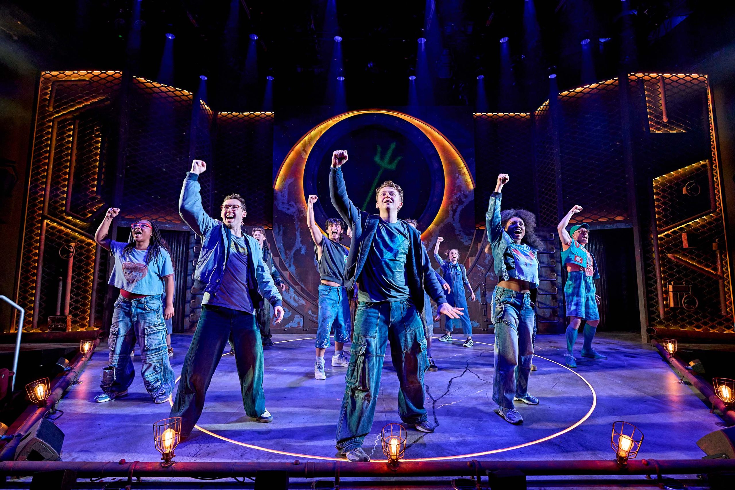 The cast of The Lightning Thief are standing on stage with their fists in the air.