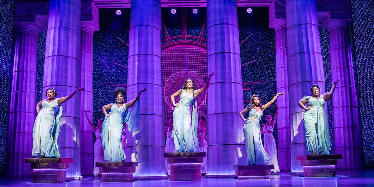 Five performers in flowing, light blue dresses pose dramatically on pedestals against a purple-lit backdrop.