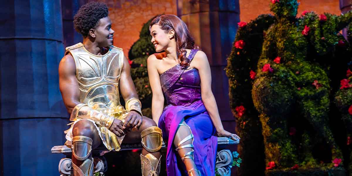 A man in golden armour and a woman in a purple dress sit together, smiling in a garden.