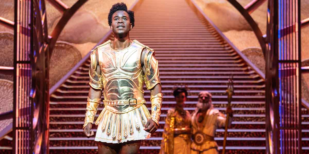 A performer in golden armour stands confidently on stage, flanked by two others in elaborate costumes.