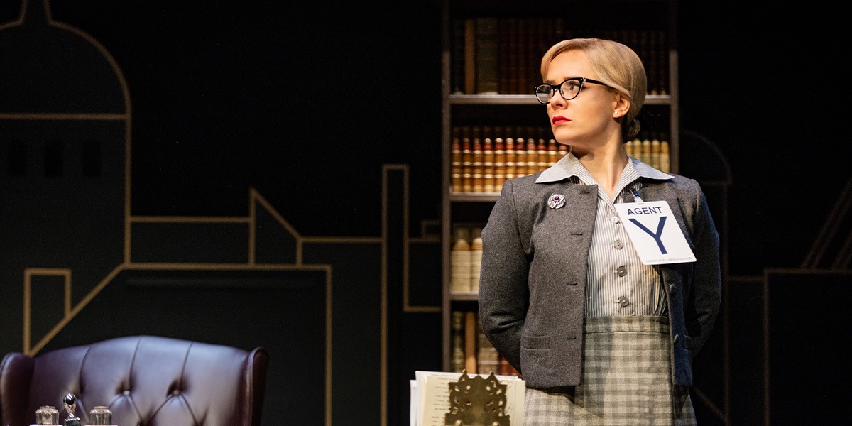 A group of actors in colourful costumes perform on stage, showcasing a comedic scene with expressive poses.