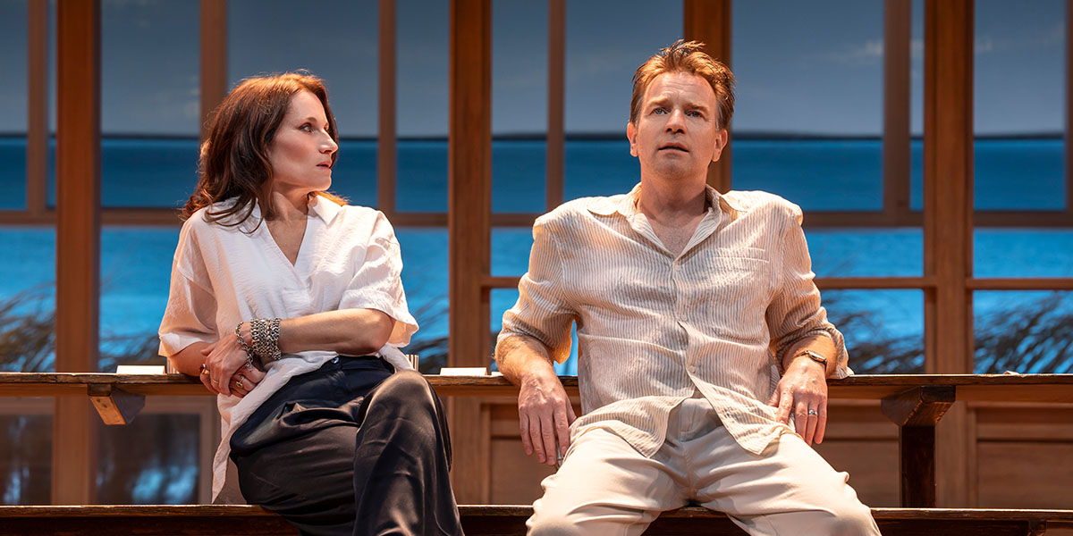 Kate Fleetwood sat on a bench, glancing at Ewan McGregor who is also sat but faces forward