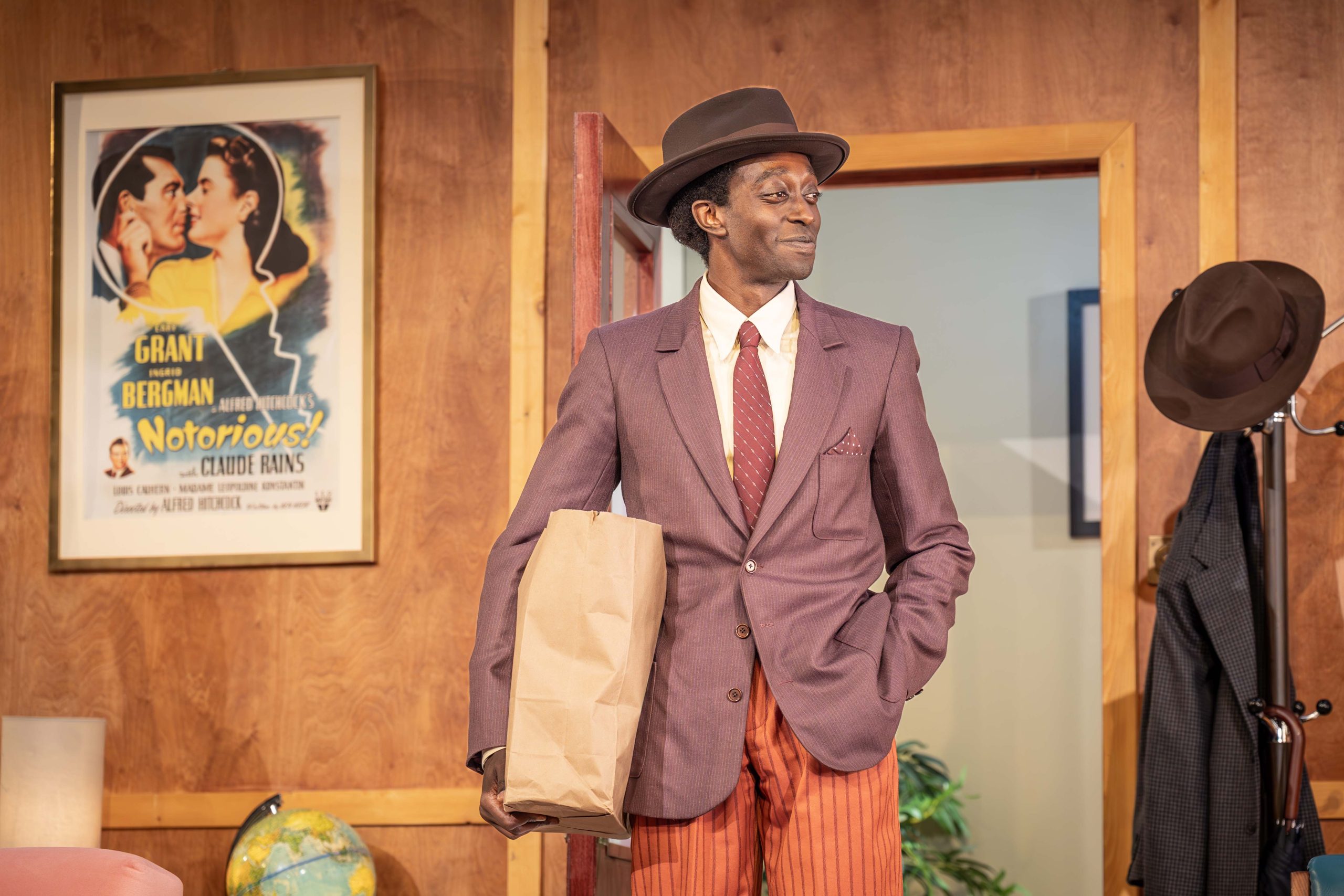 A person wearing a brown blazer, orange trousers and a brown hat is standing in front of a doorway. They are holding a brown paper bag under their right arm.