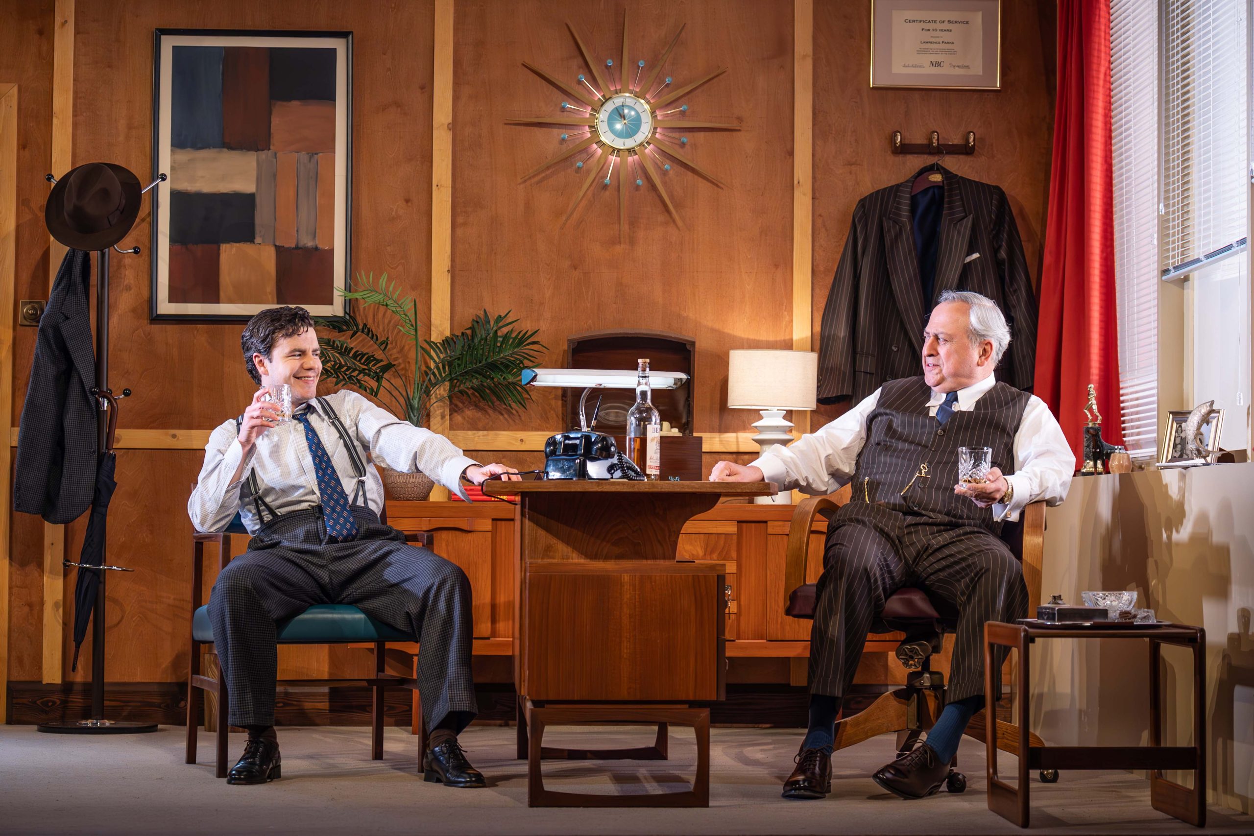 Two people are sitting at a brown desk. They are enjoying a pale brown coloured drink.