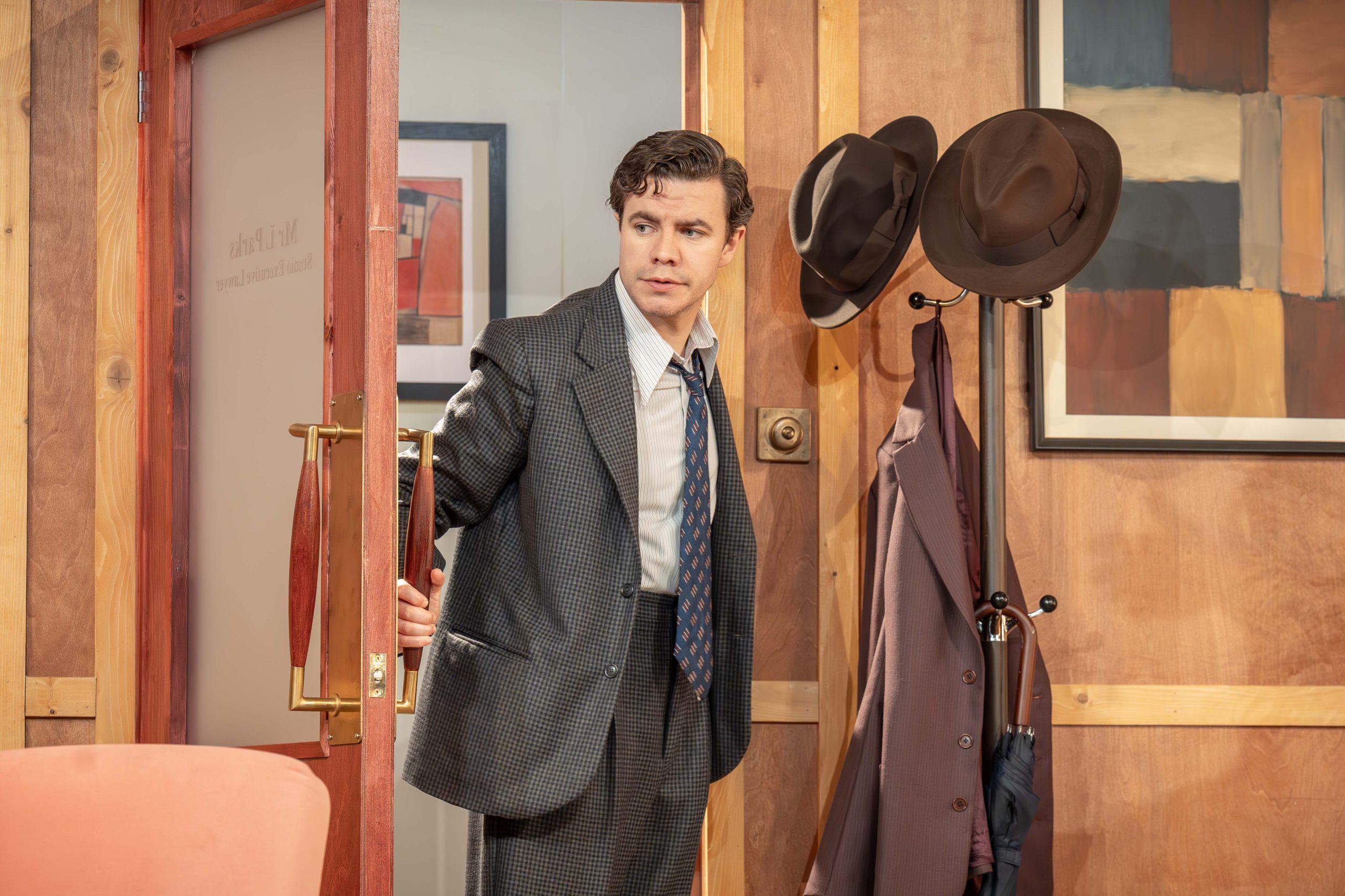 A person is standing in a doorway as if they have just entered the room. The person is wearing a dark grey suit and tie.