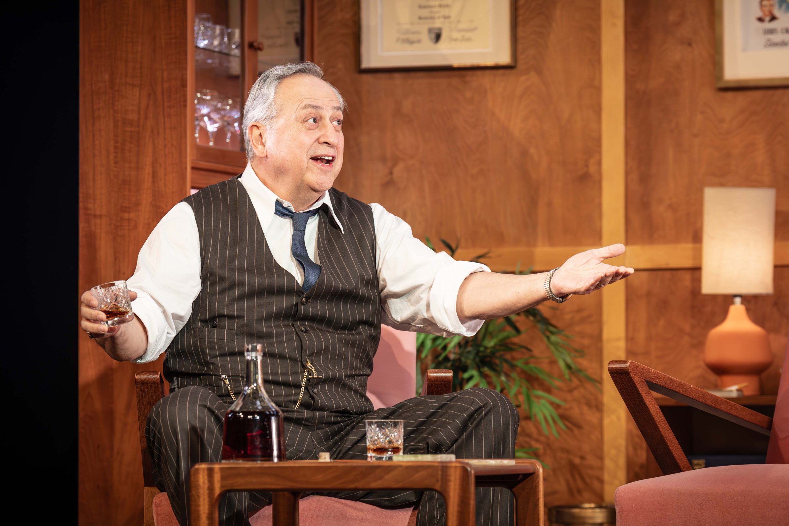 A person wearing a white shirt, brown waistcoat and tie is sitting at a small brown table. There is a bottle of brown drink in front of them.