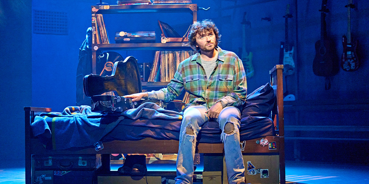 A person sits on a bunk bed surrounded by books, luggage, and a shoe rack, appearing contemplative on a stage set designed to look like a cluttered room.