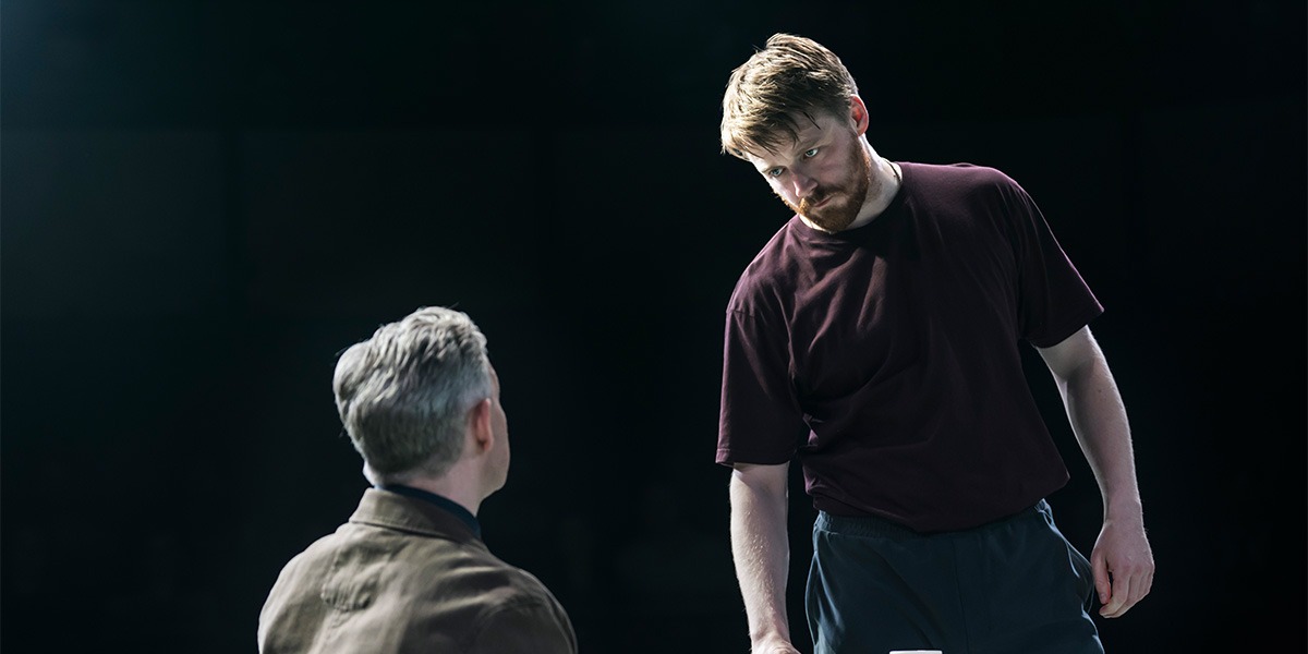 Martin Freeman (James) is sitting down, back facing the camera. Jack Lowden (Luka) is standing over them and appears unhappy.