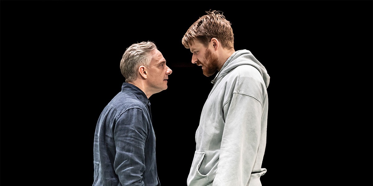 Martin Freeman and Jack Lowden sit on a wooden bench, engaged in conversation, surrounded by greenery.