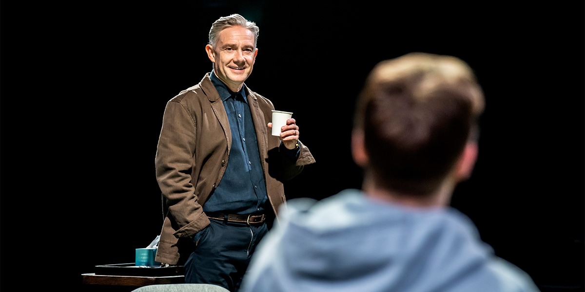 Martin Freeman (James) is standing holding a white cup and is looking towards Jack Lowden (Luka)