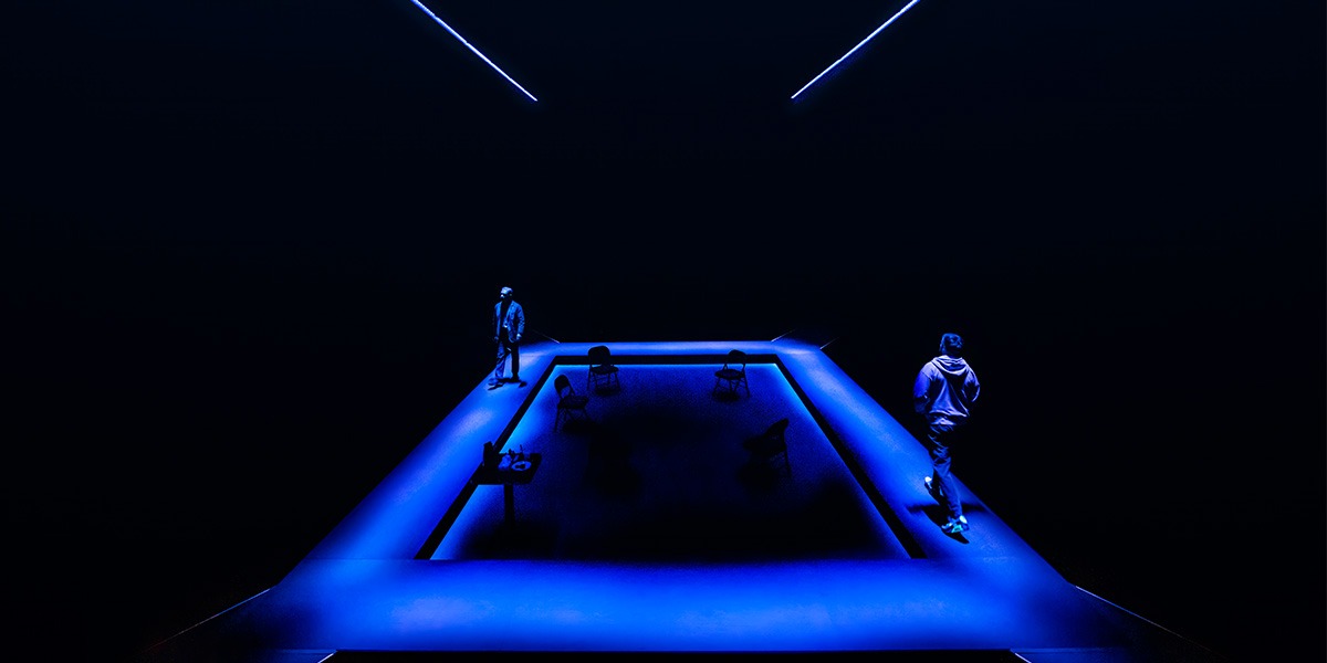Martin Freeman (James) and Jack Lowden (Luka) can be seen walking along the outside of the stage which is illuminated in blue lighting.