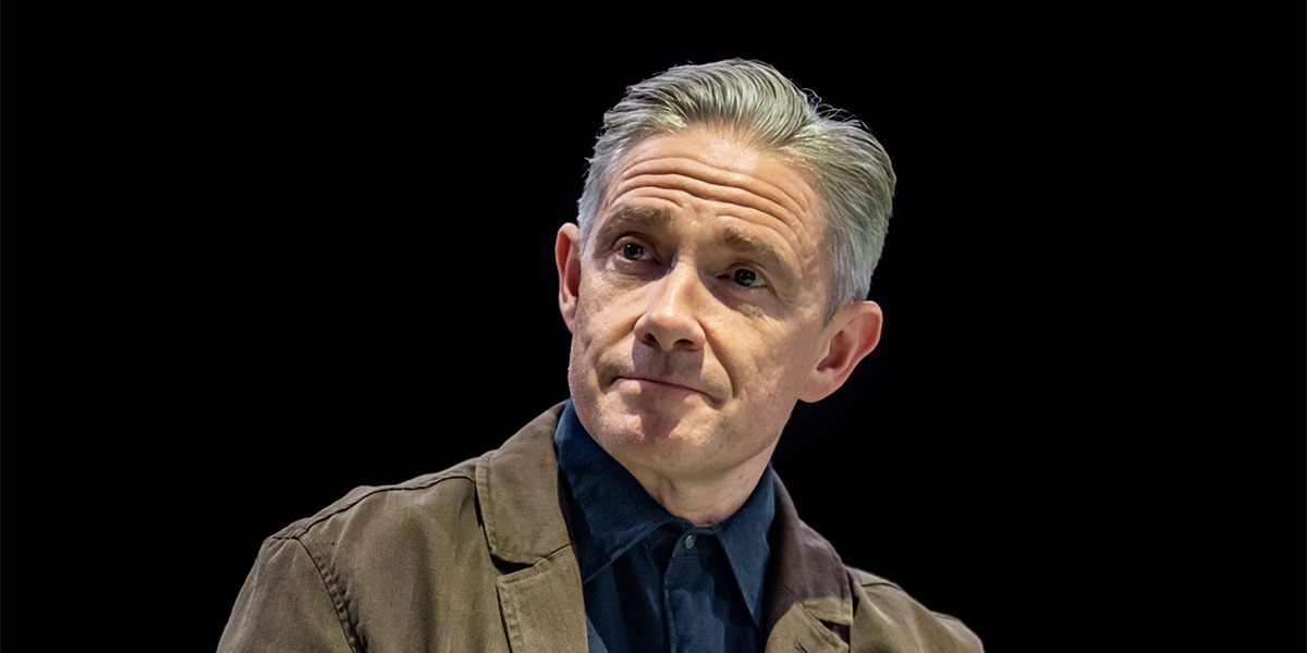 Martin Freeman (James) is sitting on stage, against a black background and appears calm.