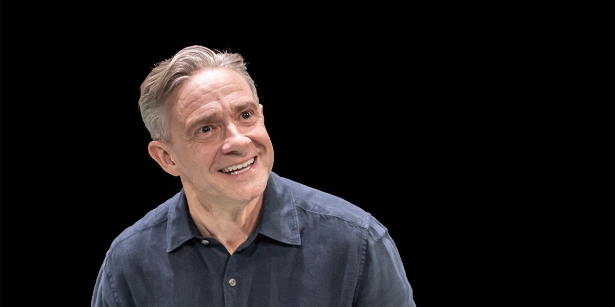 Martin Freeman (James) is sitting on stage against a black background and is smiling yet appears to be angry.