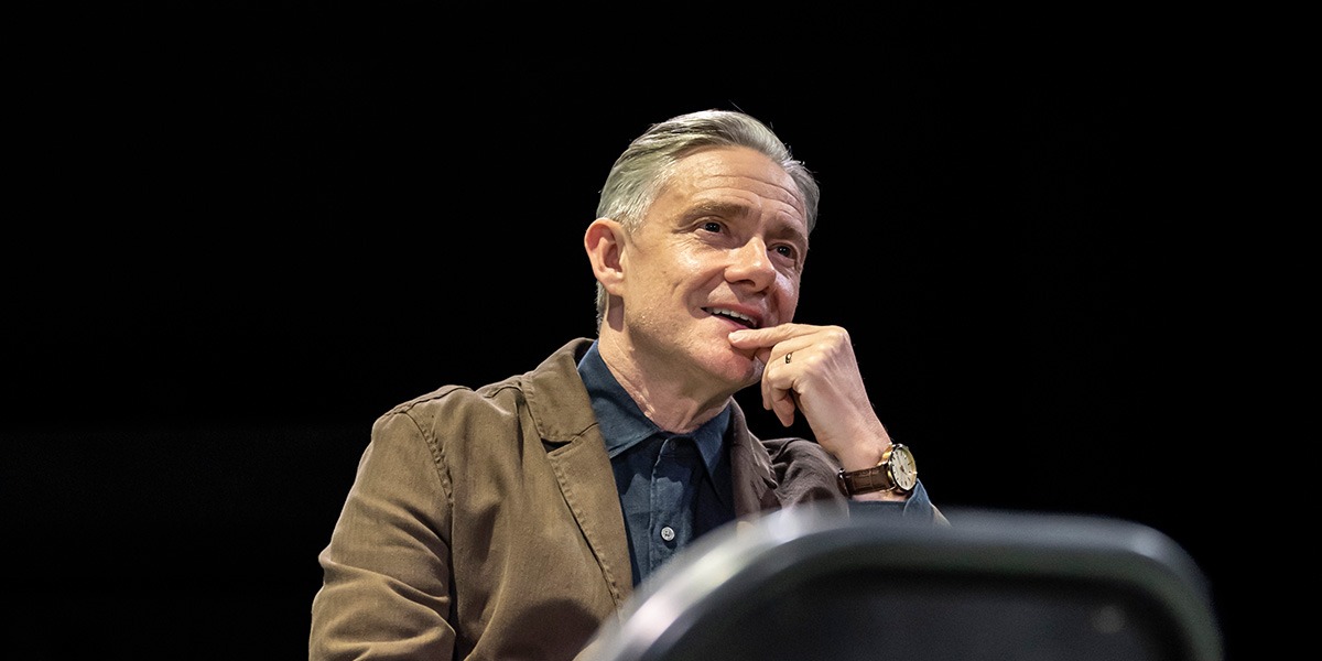 Martin Freeman (James) is sitting on stage against a black background with one hand held to the chin, as though contemplating something.
