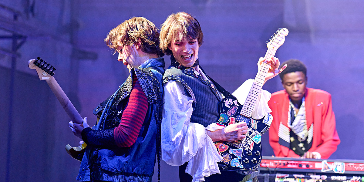 Musicians performing on stage with electric guitars, while another member plays the keyboard in the background. The setting features vibrant stage lighting.