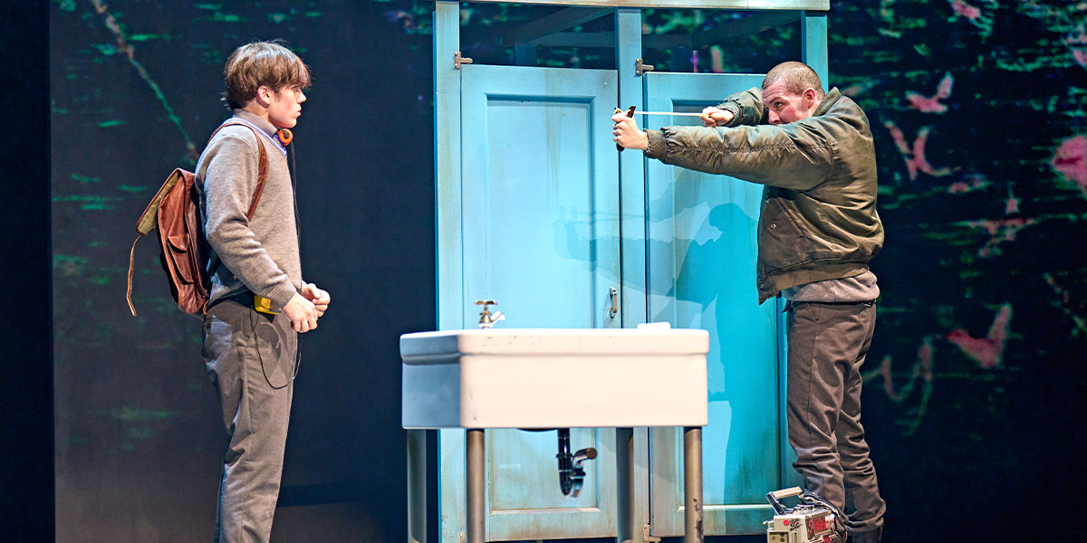 Two individuals are on stage during a performance, one standing near a sink with a backpack and the other interacting with an object by the sink.