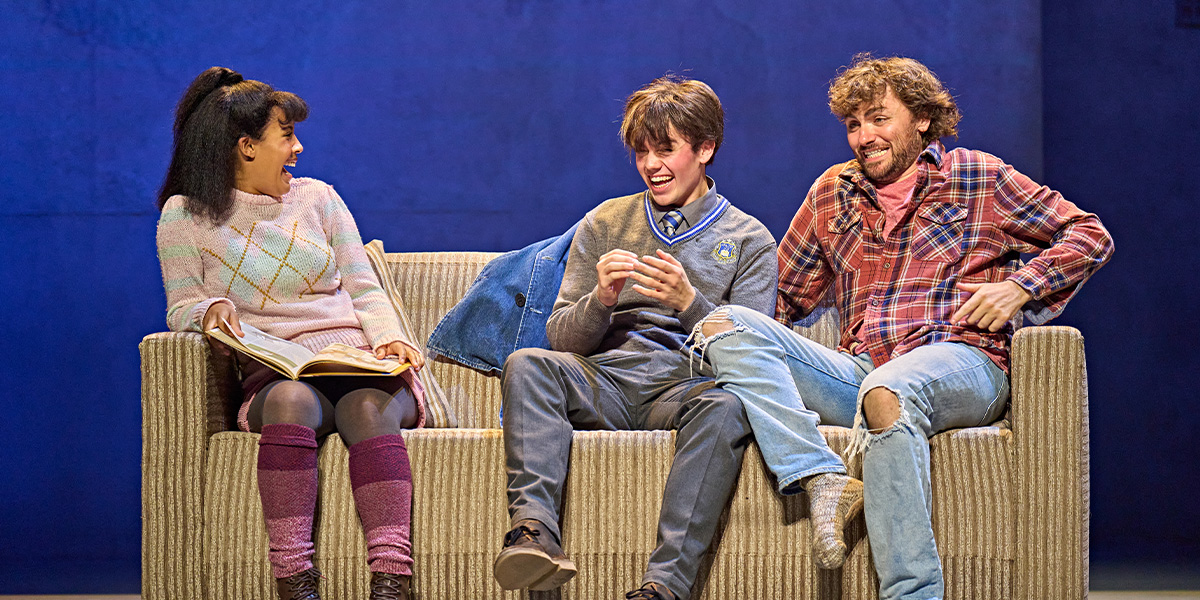Three people sitting on a sofa engaged in a lively conversation on a theatre stage, with one holding a book.