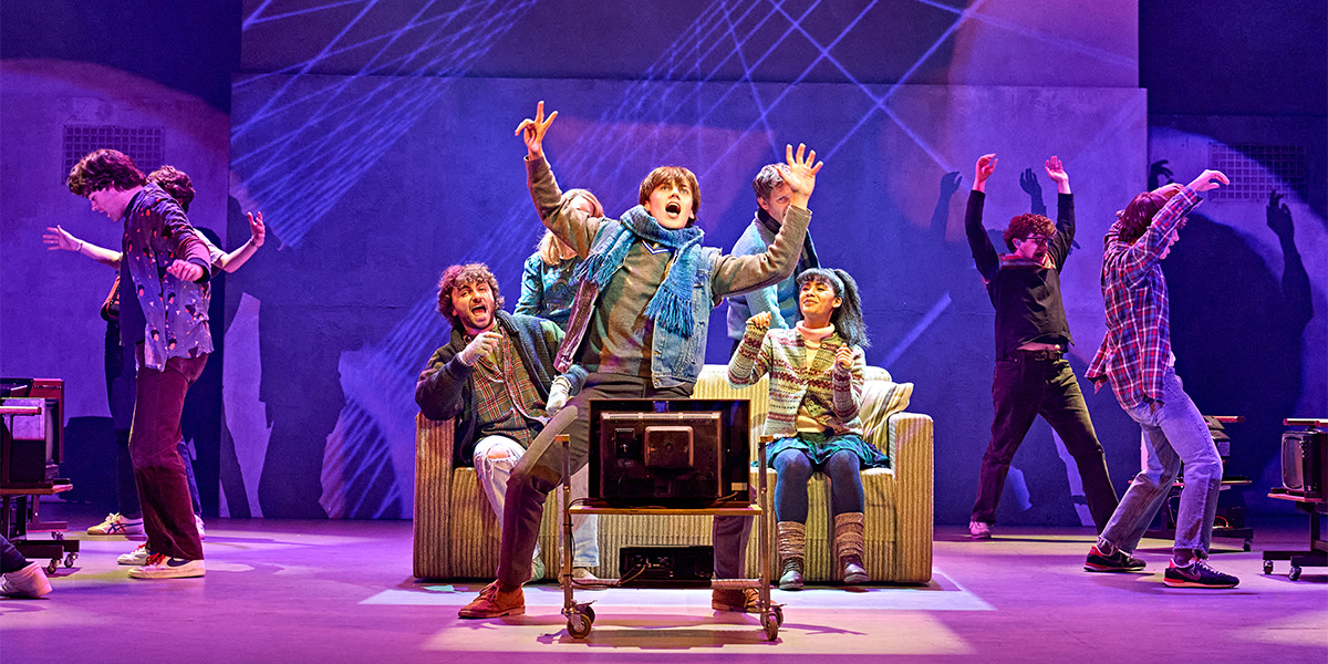 A dynamic scene from the musical "Sing Street" with five performers energetically acting on stage, with one seated in the centre in front of a television, surrounded by colourful lighting and a geometric stage design.