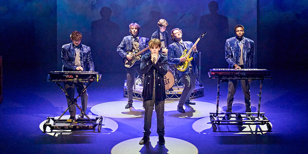 Five members of a band perform on stage, illuminated by soft blue lighting. The band members are playing keyboards, a guitar, and drums. They are dressed in matching sparkly jackets.